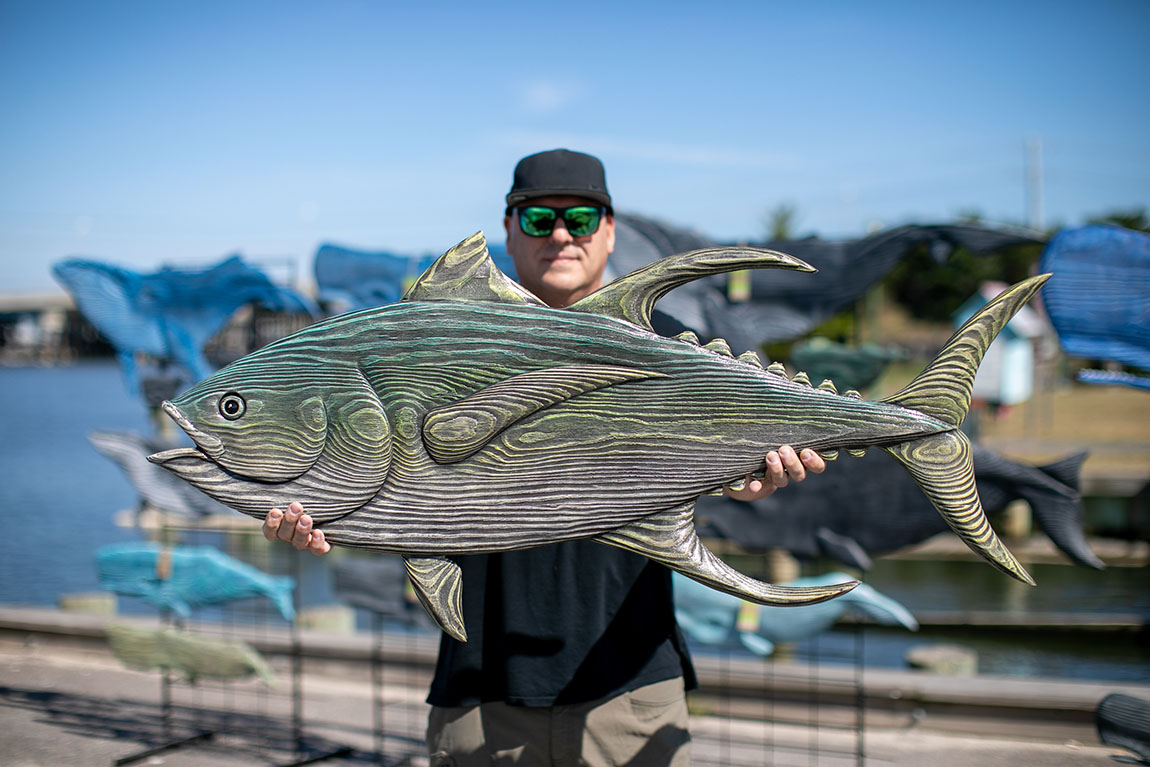 A man in a black cap and sunglasses holds a wooden carved fish out in front of him so the camera can capture the wood grain